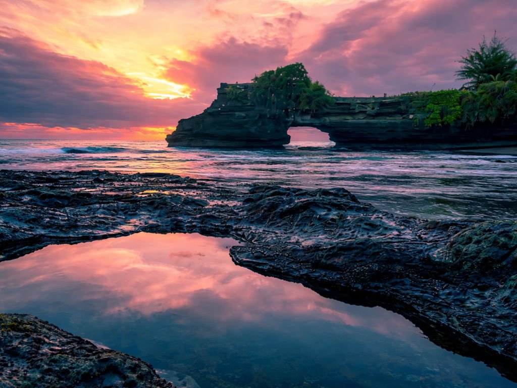 Pantai Batu Bolong saat sore hari dengan langit jingga, formasi batu karang berlubang ikonik, ombak laut, dan pantulan cahaya matahari di genangan air di antara karang.